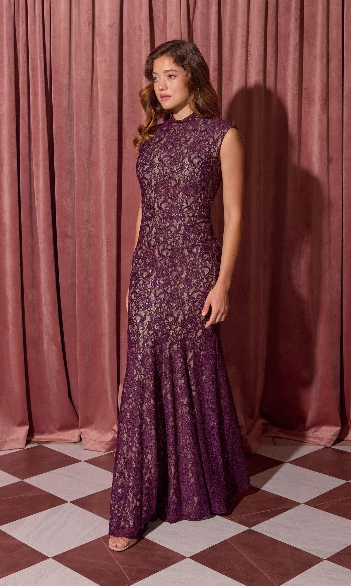 A woman stands on a checkered floor in front of mauve curtains wearing the Eureka 2061 Long Lace Formal Dress, a sleeveless purple evening gown.